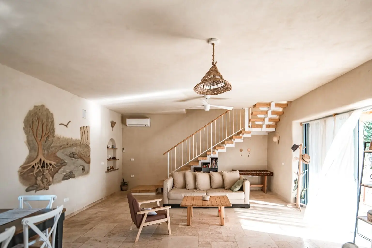 Open living room with beige sofa, wooden coffee table, staircase, and woven pendant light illuminated by sunlight from large sliding doors.